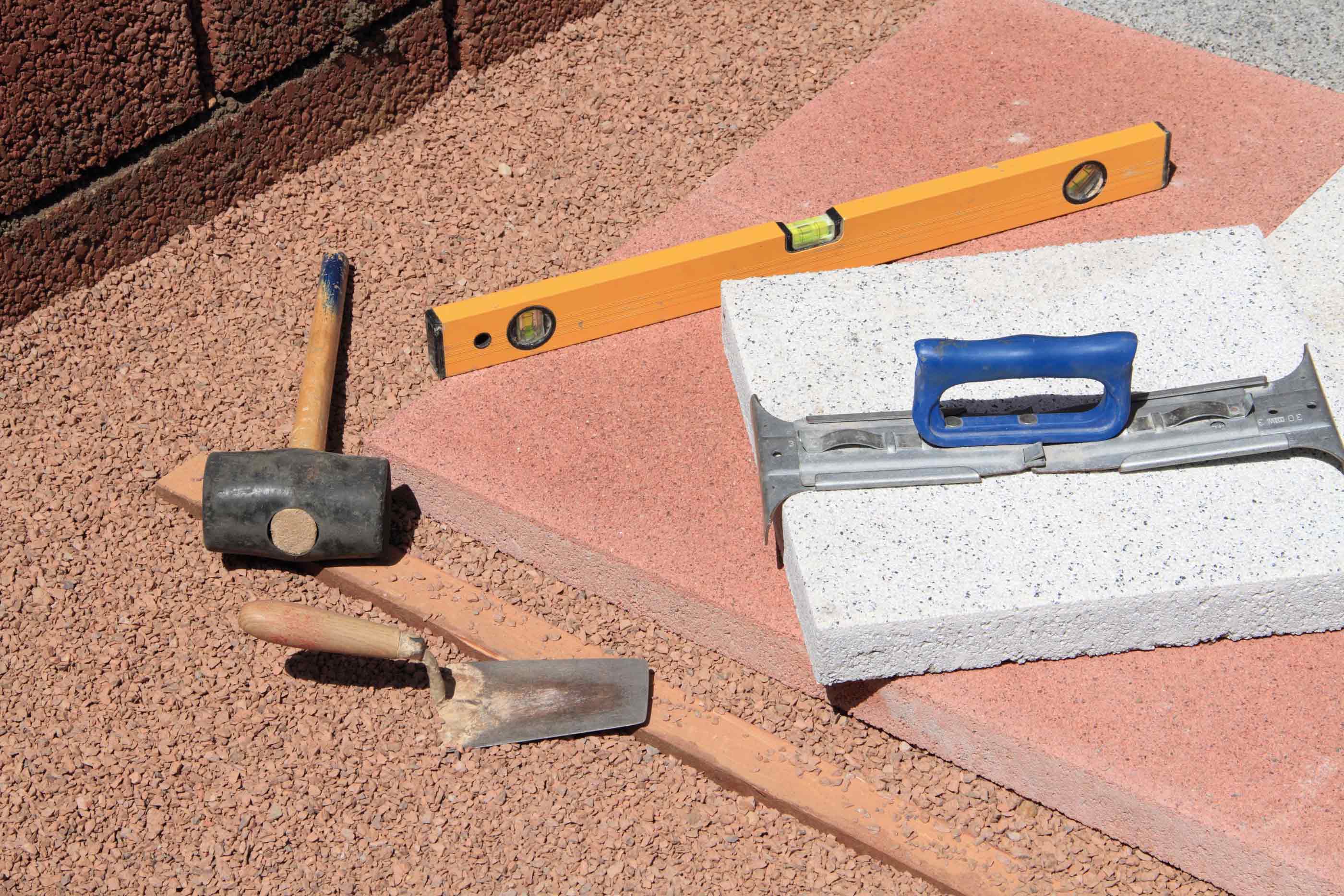 Verschiedene Zubehörteile für den Terrassenbau, ein Gummihammer, ein Plattenheber aus Metall und eine Wasserwaage werden zur genauen Verlegung eines weißen Pflastersteines verwendet.